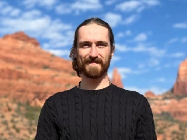 Man with a brown beard and brown hair pulled back into a bun. Wearing a black long sleeved shirt. Top half visible. The background is sandstone cliffs in Sedona, Arizona with a blue sky and scattered cloud.