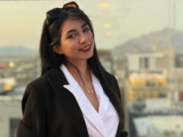 A woman wearing a black blazer and a white collared shirt underneath. Long black hair down with sunglasses on top of head. Wearing red lipstick and smiling. The background is Moonstone bar on top of the Graduate building in Tucson, Arizona and shows the Tucson sky line.