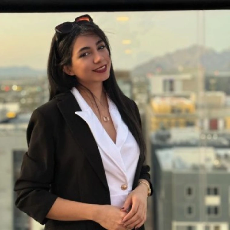 A woman wearing a black blazer and a white collared shirt underneath. Long black hair down with sunglasses on top of head. Wearing red lipstick and smiling. The background is Moonstone bar on top of the Graduate building in Tucson, Arizona and shows the Tucson sky line.